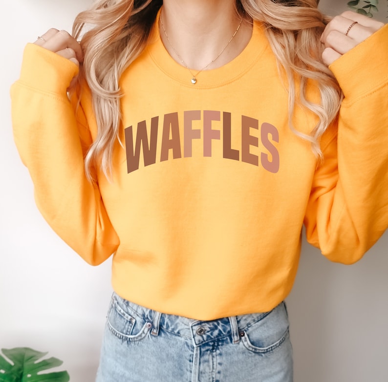 May include: A yellow sweatshirt with the word "WAFFLES" printed in brown, orange, and pink letters.