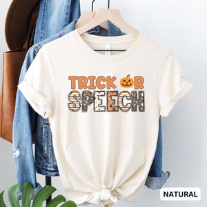 Può includere: Una t-shirt di colore naturale con il testo "TRICK OR SPEECH" in arancione, nero e bianco con una grafica di zucca.