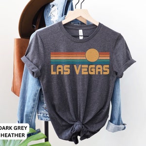 May include: Dark grey heather t-shirt with a retro-style graphic of a sunset over a striped horizon with the text "LAS VEGAS".