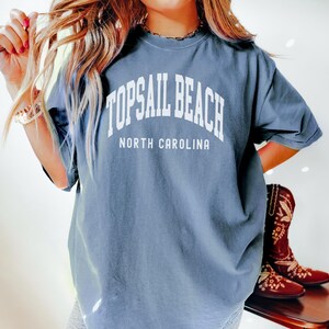 Pu&ograve; includere: T-shirt blu jeans con la scritta bianca "TOPSAIL BEACH NORTH CAROLINA". La maglietta &egrave; a maniche corte e girocollo. Sullo sfondo, stivali da cowboy marroni con ricami floreali.