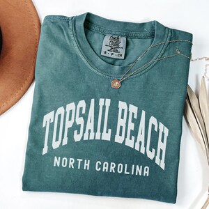 Pu&ograve; includere: Una maglietta Comfort Colors verde acqua con la scritta "TOPSAIL BEACH NORTH CAROLINA" in lettere bianche. Una collana d'oro con un ciondolo poggia sulla maglietta. Un cappello marrone e foglie di palma secche sono sullo sfondo.