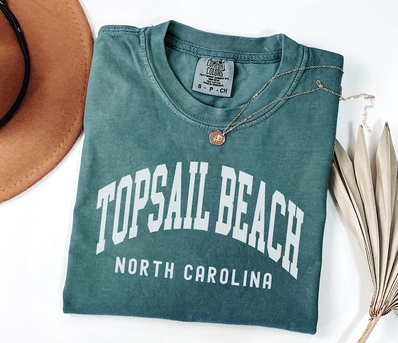 Pu&ograve; includere: Una maglietta Comfort Colors verde acqua con la scritta "TOPSAIL BEACH NORTH CAROLINA" in lettere bianche. Una collana d'oro con un ciondolo poggia sulla maglietta. Un cappello marrone e foglie di palma secche sono sullo sfondo.