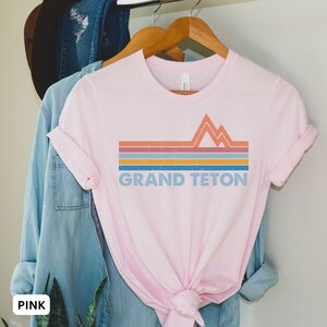 Puede incluir: Camiseta rosa con un dise&ntilde;o gr&aacute;fico retro de monta&ntilde;as y el texto "GRAND TETON" en azul.