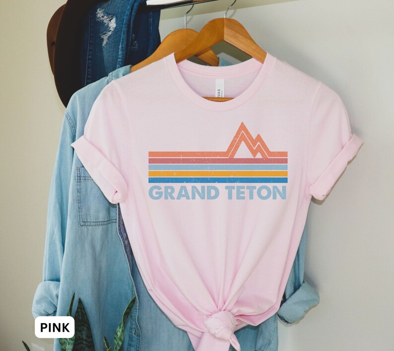 Puede incluir: Camiseta rosa con un dise&ntilde;o gr&aacute;fico retro de monta&ntilde;as y el texto "GRAND TETON" en azul.