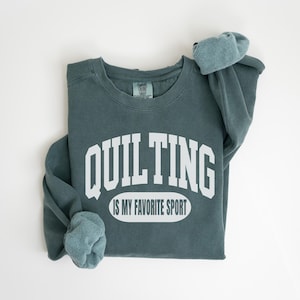 Pode incluir: Camisola verde-azulado com o texto branco "QUILTING IS MY FAVORITE SPORT". As mangas estão dobradas. A peça de roupa está dobrada sobre um fundo branco. Uma peça de roupa confortável.