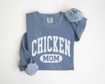 May include: A dusty blue sweatshirt with the words "CHICKEN MOM" in white block letters. The sweatshirt has a crew neck and long sleeves, with the sleeves rolled up. The Comfort Colors brand tag is visible.