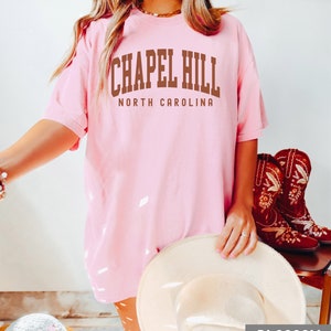 Op de afbeelding: Een roze T-shirt met de tekst "Chapel Hill North Carolina" in bruine letters.