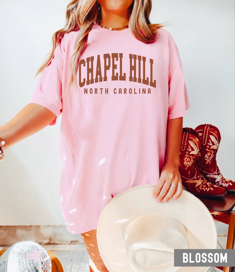 Op de afbeelding: Een roze T-shirt met de tekst "Chapel Hill North Carolina" in bruine letters.