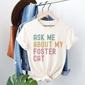 Könnte beinhalten: Cremefarbenes T-Shirt mit dem Text "ASK ME ABOUT MY FOSTER CAT" in Grün, Gold und Rosa. Das Shirt hängt an einem Holzbügel mit einer Jeansjacke und einer braunen Tragetasche im Hintergrund.