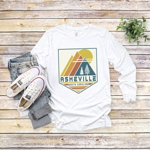May include: White long-sleeve t-shirt with a vintage-style graphic design featuring a mountain range, trees, and a sun. The graphic is in a shield shape with the text "Asheville, North Carolina" below it.