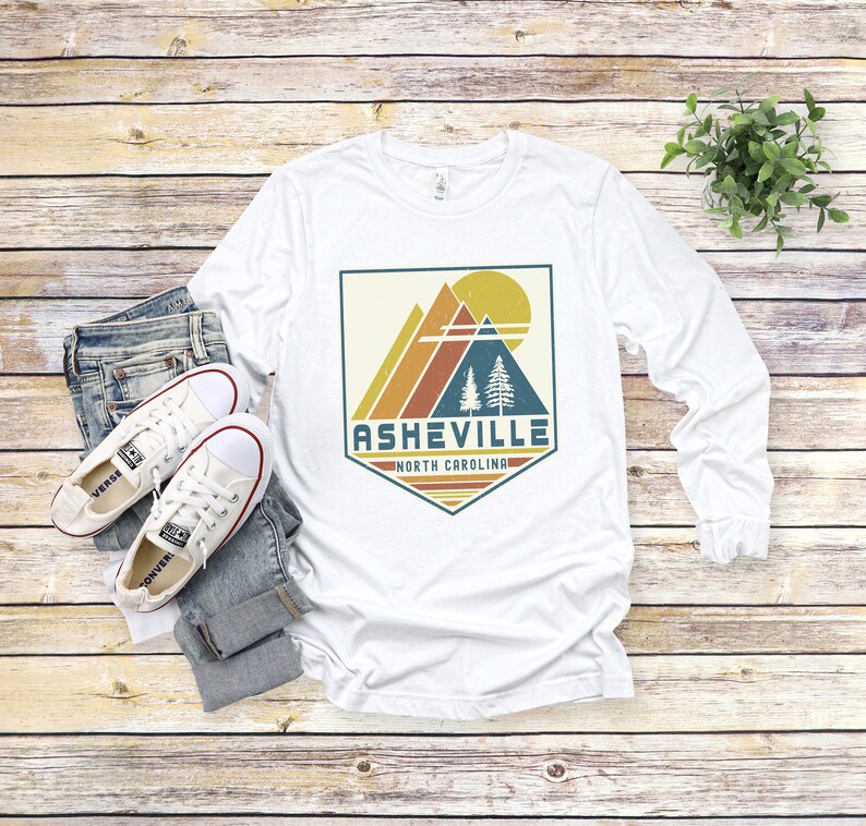 May include: White long-sleeve t-shirt with a vintage-style graphic design featuring a mountain range, trees, and a sun. The graphic is in a shield shape with the text "Asheville, North Carolina" below it.