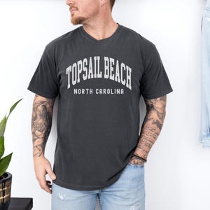 Pu&ograve; includere: T-shirt grigio scuro di Topsail Beach, Carolina del Nord. La maglietta presenta un testo bianco ad arco che recita "TOPSAIL BEACH" sopra "NORTH CAROLINA". La maglietta &egrave; girocollo ed &egrave; indossata da una persona.