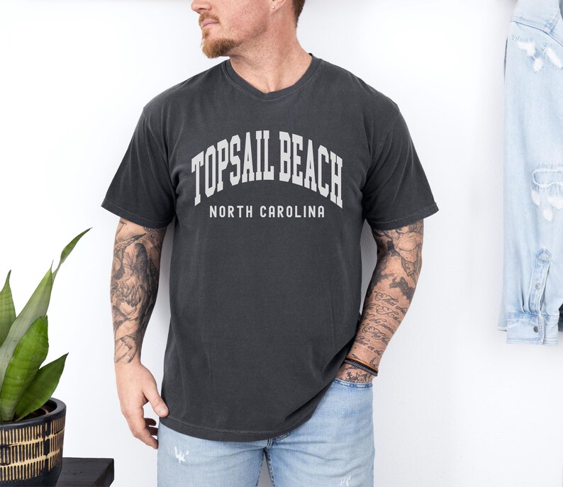 Pu&ograve; includere: T-shirt grigio scuro di Topsail Beach, Carolina del Nord. La maglietta presenta un testo bianco ad arco che recita "TOPSAIL BEACH" sopra "NORTH CAROLINA". La maglietta &egrave; girocollo ed &egrave; indossata da una persona.