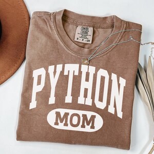 May include: A brown t-shirt with the words "PYTHON MOM" in white block letters. The shirt has a crew neck and a small tag at the neckline. A silver necklace with a pendant is draped across the shirt.