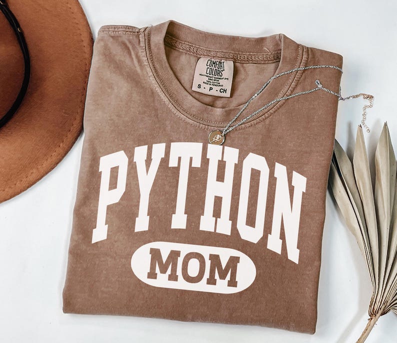 May include: A brown t-shirt with the words "PYTHON MOM" in white block letters. The shirt has a crew neck and a small tag at the neckline. A silver necklace with a pendant is draped across the shirt.