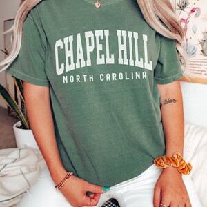 Op de afbeelding: Een groen T-shirt met de tekst "Chapel Hill North Carolina" in wit. Het shirt wordt gedragen door een persoon met lang blond haar en een gouden ketting.