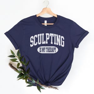 K&ouml;nnte beinhalten: Marineblaues T-Shirt mit dem wei&szlig;en Text "SCULPTING IS MY THERAPY". Der Text ist &uuml;ber einem wei&szlig;en Ovalbogen angeordnet. Das T-Shirt h&auml;ngt an einem Holzb&uuml;gel vor wei&szlig;em Hintergrund. Unten ist ein Zweig mit Gr&uuml;n.