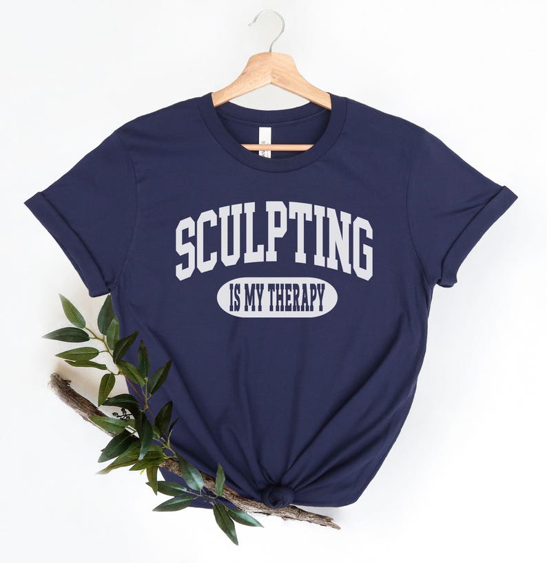 K&ouml;nnte beinhalten: Marineblaues T-Shirt mit dem wei&szlig;en Text "SCULPTING IS MY THERAPY". Der Text ist &uuml;ber einem wei&szlig;en Ovalbogen angeordnet. Das T-Shirt h&auml;ngt an einem Holzb&uuml;gel vor wei&szlig;em Hintergrund. Unten ist ein Zweig mit Gr&uuml;n.