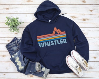 May include: A navy blue hoodie with a vintage-style graphic of a mountain range and the word "WHISTLER" in white text. The hoodie is laid out on a white wooden surface with a pair of white sneakers.