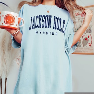 May include: A light blue oversized t-shirt with the text "JACKSON HOLE WYOMING" in navy blue. The shirt is made of a soft, comfortable fabric and has a relaxed fit. The shirt is shown on a model wearing it with a casual look.
