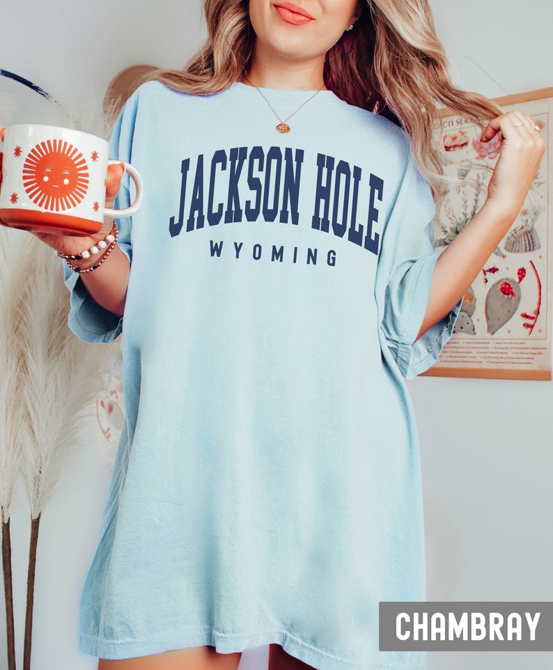 May include: A light blue oversized t-shirt with the text "JACKSON HOLE WYOMING" in navy blue. The shirt is made of a soft, comfortable fabric and has a relaxed fit. The shirt is shown on a model wearing it with a casual look.