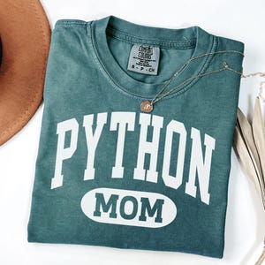 May include: A teal Comfort Colors t-shirt with the words "PYTHON MOM" in white block letters. A gold necklace with a circular pendant rests on the shirt. A brown hat and dried palm leaf are in the background.