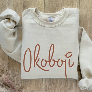 May include: A beige sweatshirt with the word "Okoboji" written in brown cursive lettering.