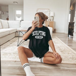 K&ouml;nnte beinhalten: Schwarzes T-Shirt mit dem wei&szlig;en Text "SCULPTING IS MY THERAPY", getragen mit wei&szlig;en Shorts und Socken. Die Socken haben schwarze Streifen oben. Die Person sitzt auf einem Teppich in einem Wohnzimmer.