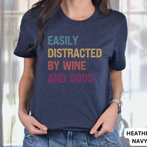 May include: A heather navy blue t-shirt with the text "EASILY DISTRACTED BY WINE AND DOGS" in a vintage style font.