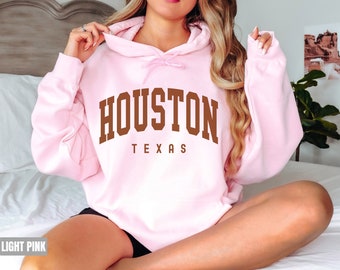 Könnte beinhalten: Ein hellrosa Hoodie mit dem Text "HOUSTON TEXAS" in braunen Buchstaben.