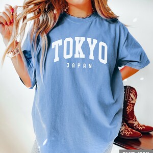 Pode incluir: Uma camiseta azul oversized com o texto "TOKYO JAPAN" em branco. A camiseta est&aacute; sendo usada por uma pessoa com cabelo loiro comprido. A pessoa est&aacute; usando shorts jeans azuis e botas de cowboy marrons.