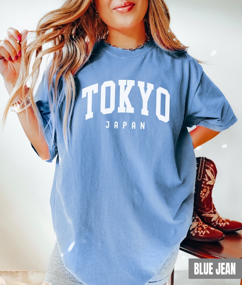 Pode incluir: Uma camiseta azul oversized com o texto "TOKYO JAPAN" em branco. A camiseta est&aacute; sendo usada por uma pessoa com cabelo loiro comprido. A pessoa est&aacute; usando shorts jeans azuis e botas de cowboy marrons.