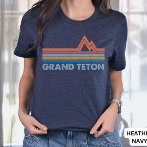 Puede incluir: Una camiseta azul marino jaspeada con un gr&aacute;fico retro de monta&ntilde;as y el texto "GRAND TETON" en azul claro.