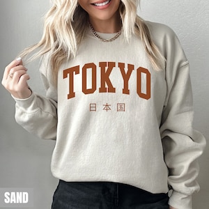 May include: A sand-colored sweatshirt with the word "TOKYO" printed in brown on the front. The Japanese characters for "Japan" are printed below the word "TOKYO".