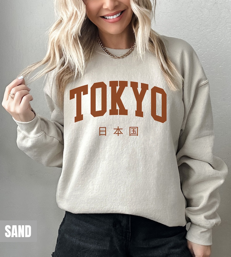 May include: A sand-colored sweatshirt with the word "TOKYO" printed in brown on the front. The Japanese characters for "Japan" are printed below the word "TOKYO".