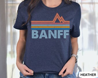 May include: A heather navy blue t-shirt with a vintage-style graphic of mountains and the word "BANFF" in a faded blue color.