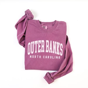 May include: A mauve-colored long-sleeved sweatshirt with the words "OUTER BANKS" and "NORTH CAROLINA" printed in white block letters. The sweatshirt is folded, with the sleeves bunched up. The tag on the collar reads "Comfort Colors".