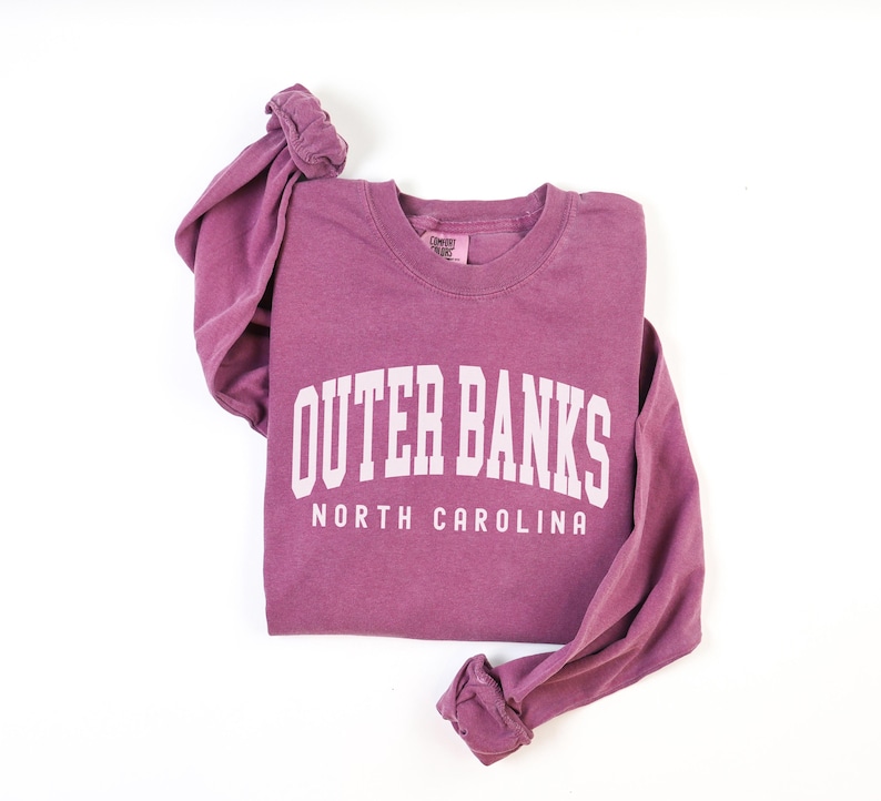 May include: A mauve-colored long-sleeved sweatshirt with the words "OUTER BANKS" and "NORTH CAROLINA" printed in white block letters. The sweatshirt is folded, with the sleeves bunched up. The tag on the collar reads "Comfort Colors".