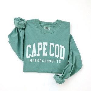 May include: A seafoam green long-sleeve shirt with the words "CAPE COD MASSACHUSETTS" printed in white block letters. The shirt is folded, with the sleeves rolled up. The shirt is made of a soft, comfortable material.