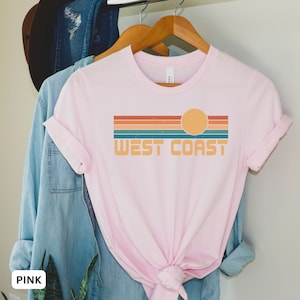 May include: A pink t-shirt with a retro-style graphic design featuring a sunset and the text "WEST COAST".