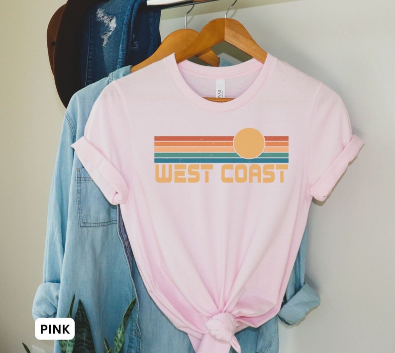 May include: A pink t-shirt with a retro-style graphic design featuring a sunset and the text "WEST COAST".