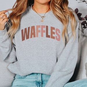 May include: A gray sweatshirt with the word "WAFFLES" printed in a rainbow of colors across the chest.