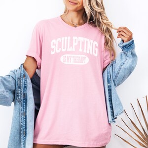 K&ouml;nnte beinhalten: Hellrosa T-Shirt mit wei&szlig;em Aufdruck "SCULPTING IS MY THERAPY". Dar&uuml;ber eine hellblaue Jeansjacke. Der Look wirkt entspannt und l&auml;ssig.