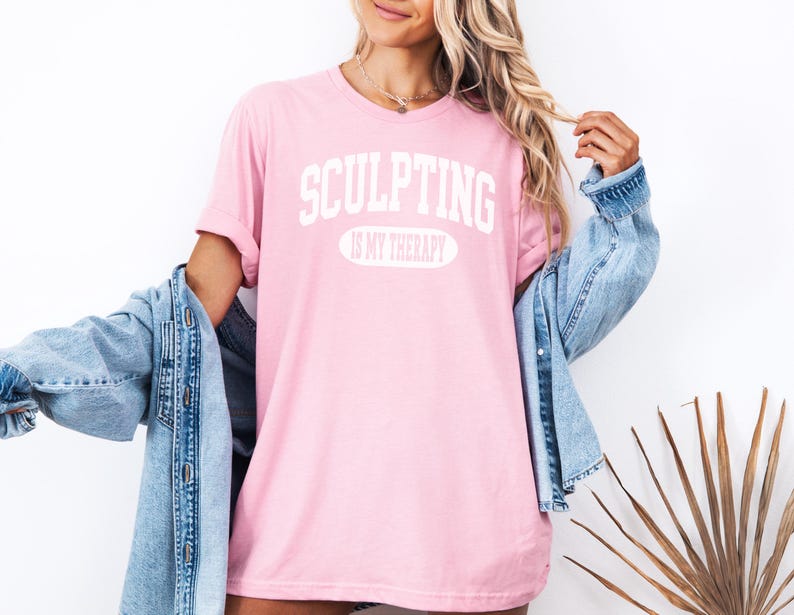 K&ouml;nnte beinhalten: Hellrosa T-Shirt mit wei&szlig;em Aufdruck "SCULPTING IS MY THERAPY". Dar&uuml;ber eine hellblaue Jeansjacke. Der Look wirkt entspannt und l&auml;ssig.
