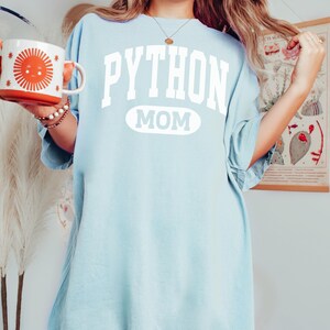 May include: Pale blue t-shirt featuring the text "PYTHON MOM" in white. The word "PYTHON" is above a white oval. The person is holding a white and orange mug with a sun design.
