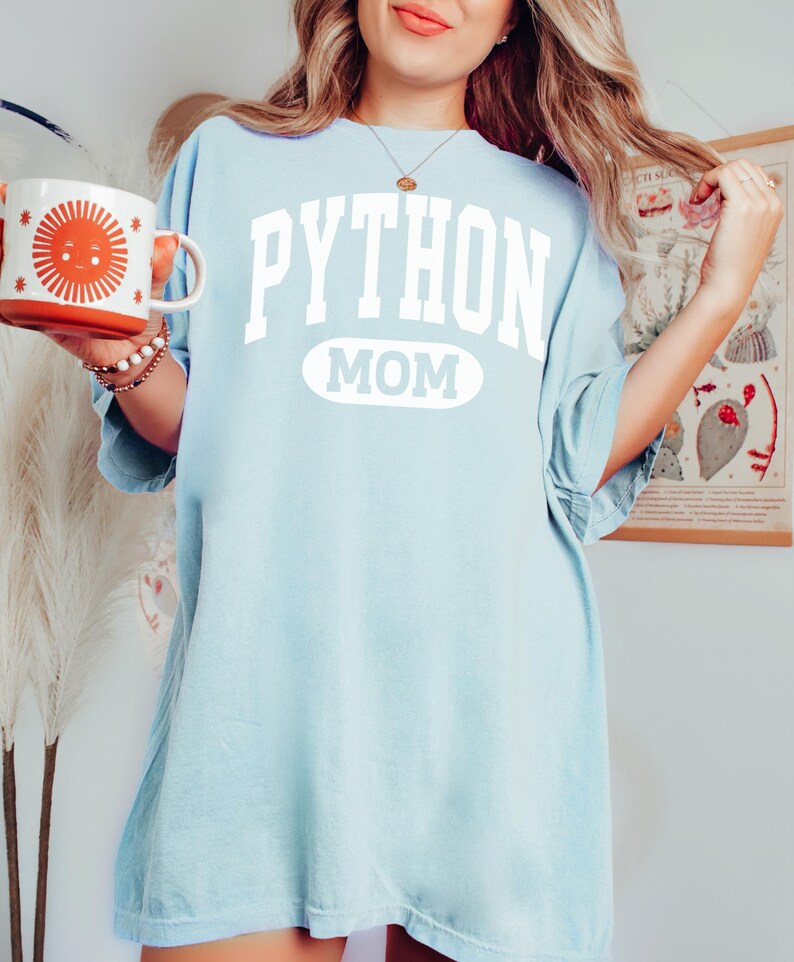 May include: Pale blue t-shirt featuring the text "PYTHON MOM" in white. The word "PYTHON" is above a white oval. The person is holding a white and orange mug with a sun design.