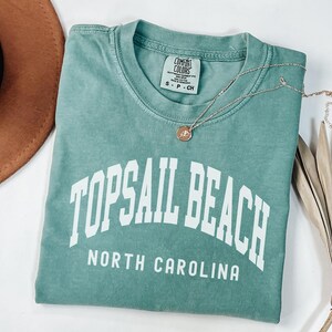 Pu&ograve; includere: Una maglietta verde acqua con la scritta "TOPSAIL BEACH NORTH CAROLINA" a lettere bianche. Una collana d'argento con un piccolo ciondolo poggia sulla maglietta. Un cappello marrone &egrave; visibile nell'angolo in alto a sinistra.