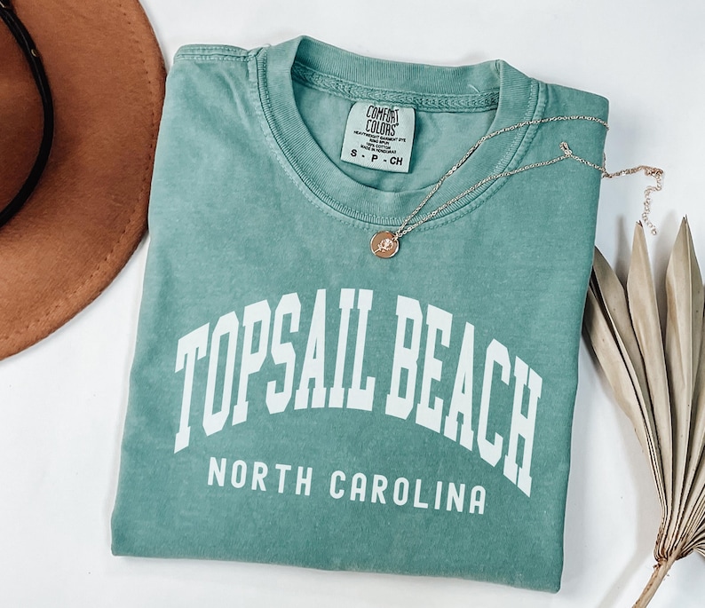 Pu&ograve; includere: Una maglietta verde acqua con la scritta "TOPSAIL BEACH NORTH CAROLINA" a lettere bianche. Una collana d'argento con un piccolo ciondolo poggia sulla maglietta. Un cappello marrone &egrave; visibile nell'angolo in alto a sinistra.
