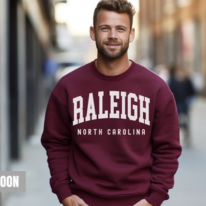 Pode incluir: Camisola de cor vinho com texto branco que diz "Raleigh North Carolina".