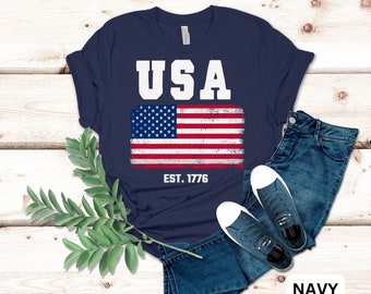 May include: A navy blue t-shirt with a white distressed American flag graphic and the text "USA EST. 1776".